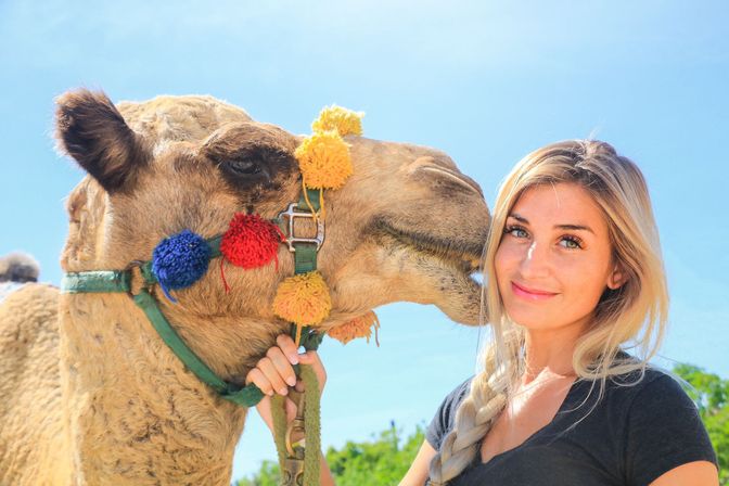 Smiling woman with braided hair poses with a decorated camel adorned with colorful pom-poms under a bright blue sky