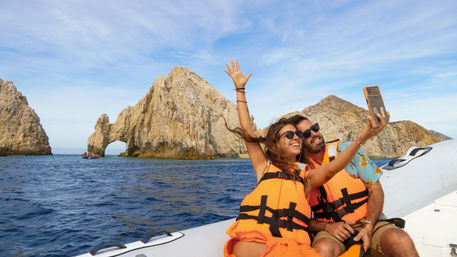 Two people in bright orange life jackets take a selfie on a small boat off Cabo San Lucas, Mexico, with the iconic El Arco rock arch rising from the blue ocean under a sunny sky
