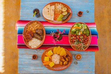 Colorful Mexican-style brunch spread on a rustic blue table with a serape runner: clay plates of tortillas, beans, rice, plantains, corn, nachos and salad.