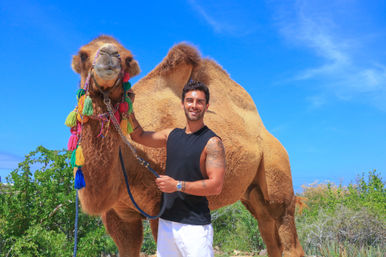 Smiling man in a sleeveless black top and white shorts holding a large camel decorated with colorful tassels, standing under a vivid blue sky with scrubby desert vegetation.