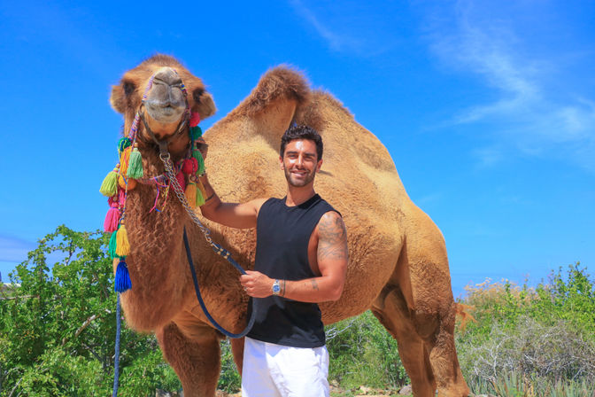 Smiling man in a sleeveless black top and white shorts holding a large camel decorated with colorful tassels, standing under a vivid blue sky with scrubby desert vegetation.