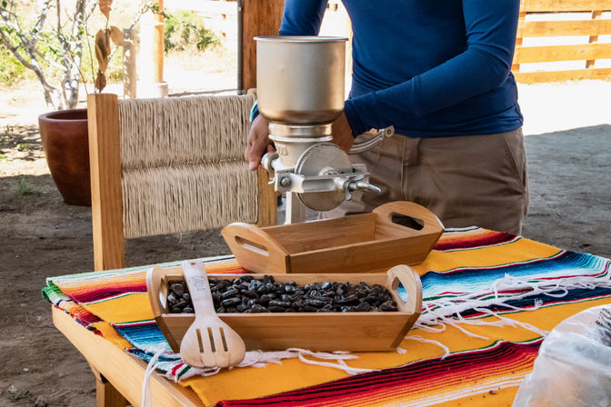 Hand-crank coffee grinder held by a person in a blue shirt above wooden serving trays of dark roasted coffee beans on a colorful woven serape covering a rustic outdoor table.