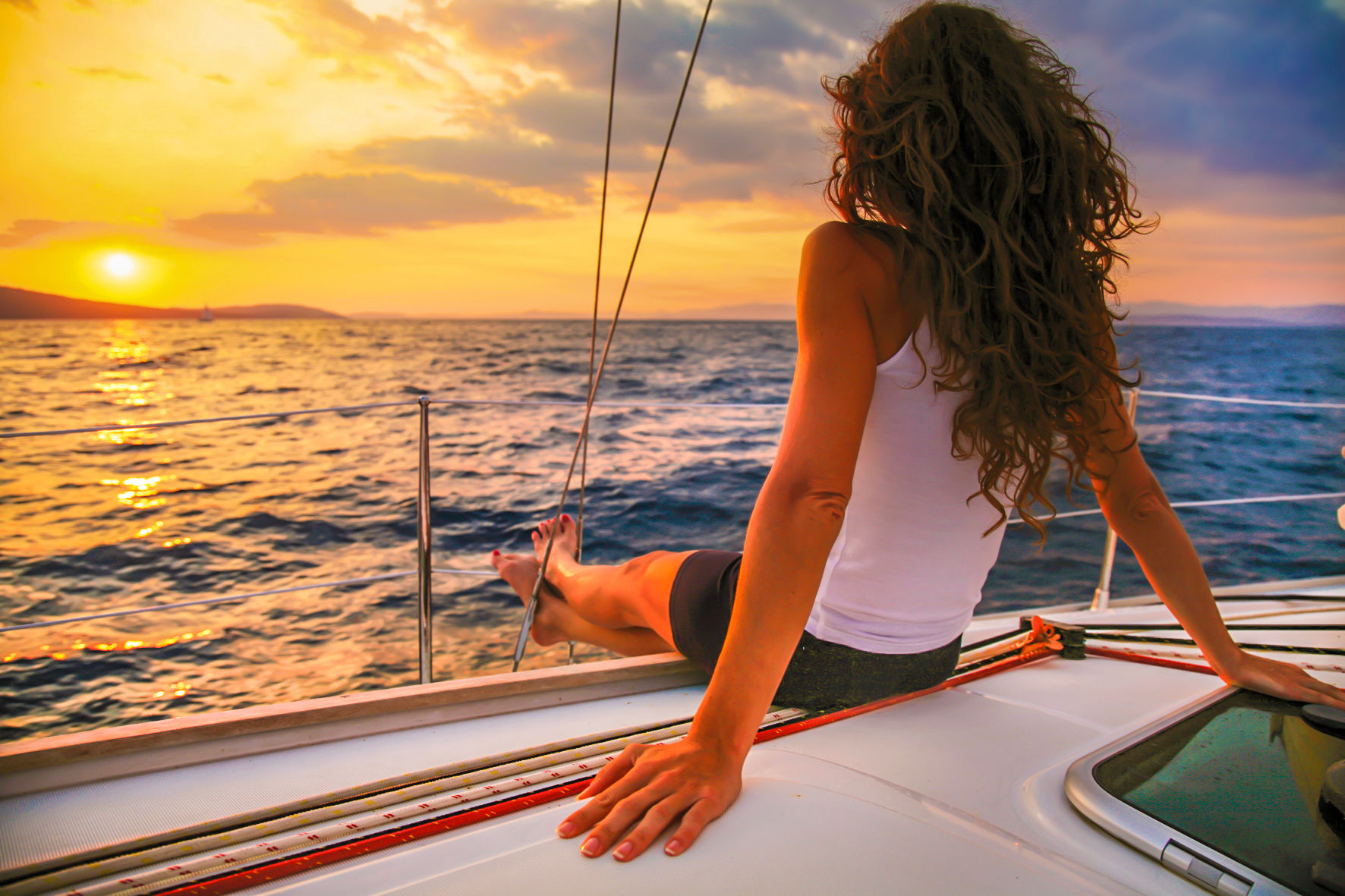 Woman lounging on a sailboat deck during a golden sunset, legs over the railing and gazing at the sparkling ocean horizon