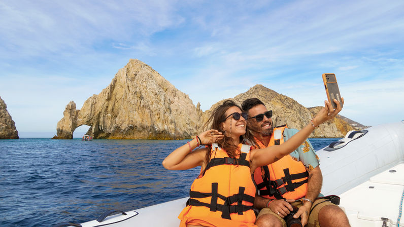 Smiling pair wearing orange life jackets and sunglasses take a selfie on an inflatable boat with blue ocean and the iconic rock arch of Cabo San Lucas rising from the sea in the background.
