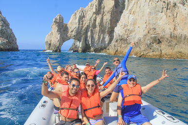 Group of cheerful people wearing bright orange life jackets on an inflatable boat, posing and waving near a dramatic coastal rock arch and turquoise sea under a clear blue sky.