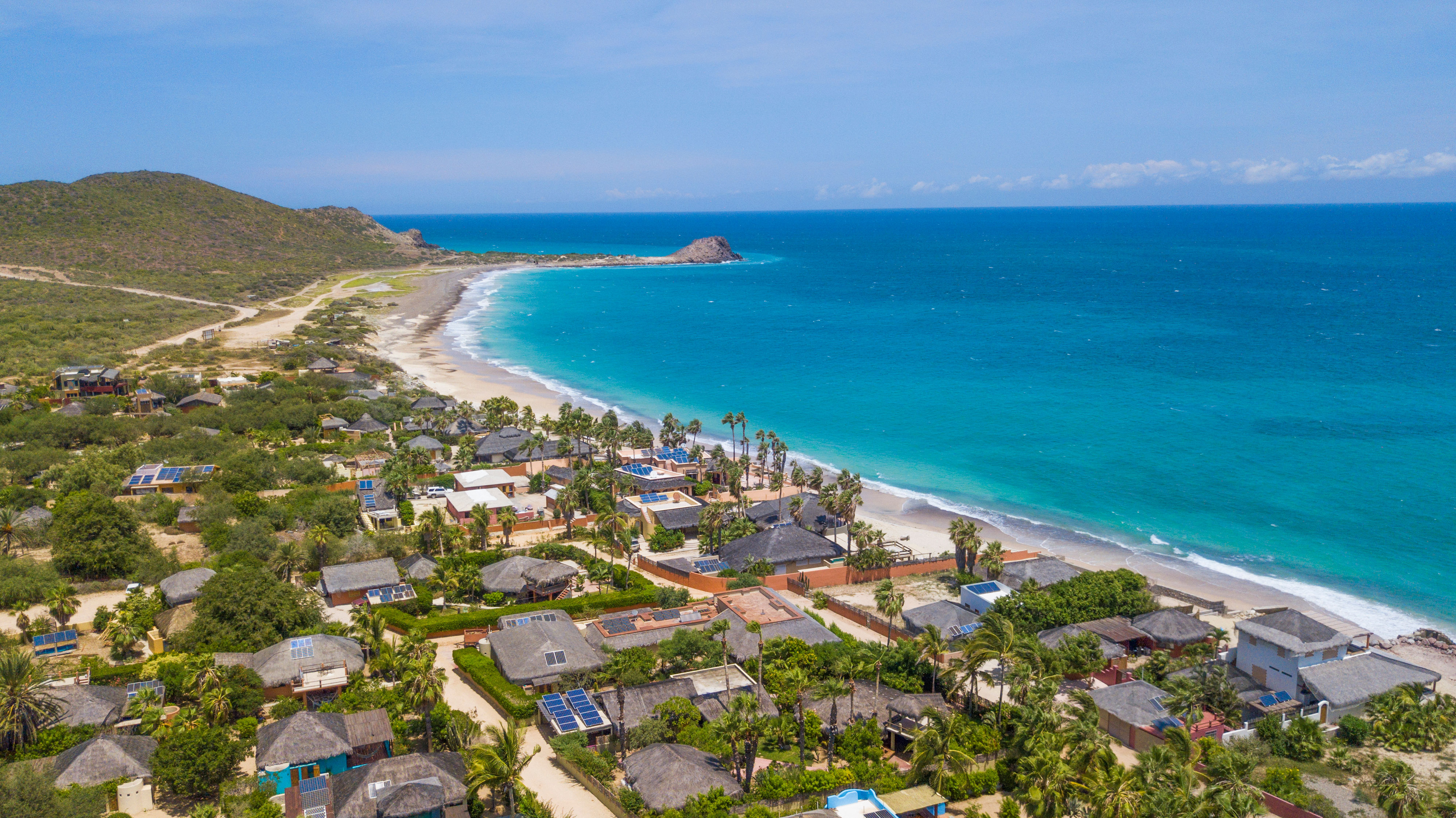 Aerial view of a sun-drenched tropical coastline with turquoise water, a crescent sandy beach, palm‑fringed beachfront villas and low green hills