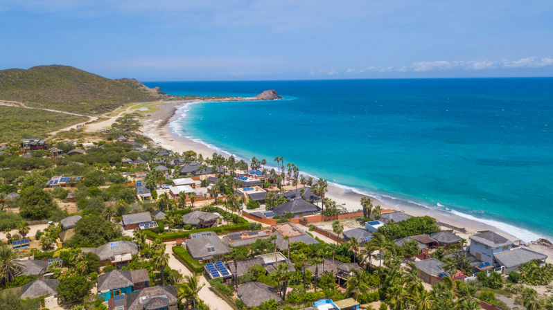 Aerial view of a sun-drenched tropical coastline with turquoise water, a crescent sandy beach, palm‑fringed beachfront villas and low green hills