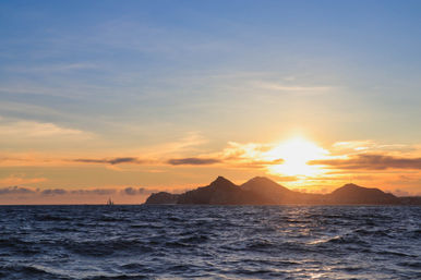 Golden sunset over choppy ocean waves with a lone sailboat and silhouetted coastal mountains on the horizon.