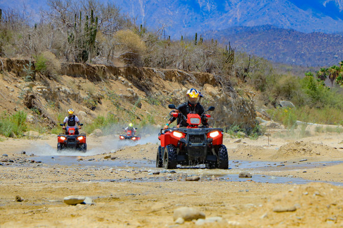 Three riders on red ATVs splashing through a shallow desert stream in a rocky canyon with cacti and distant blue mountains