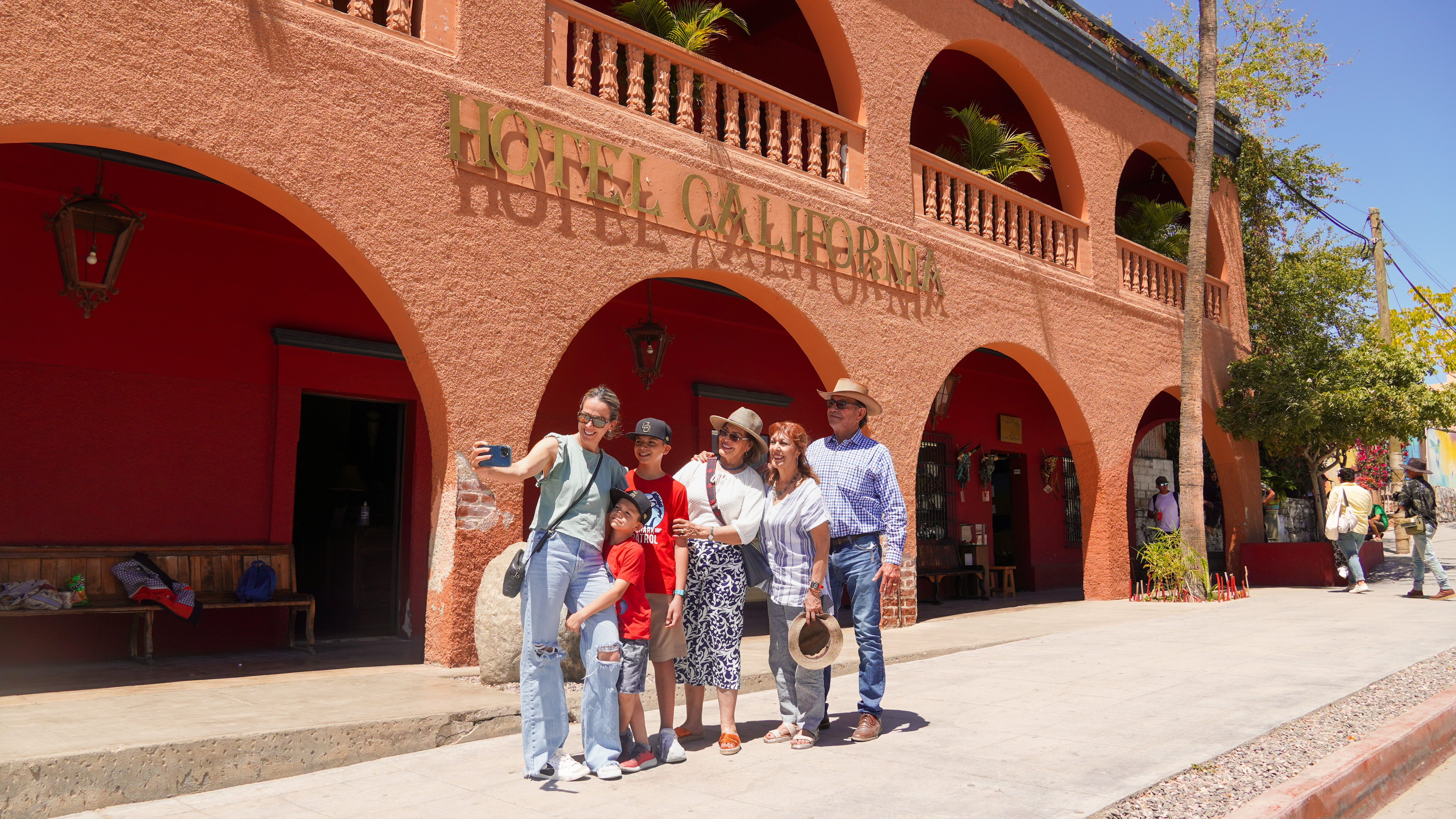 Multigenerational family taking a selfie outside an orange stucco building with arched balconies in a sunny Mexican town plaza