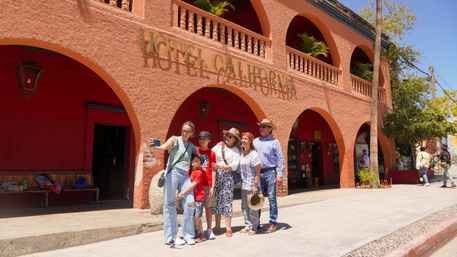 Multigenerational family taking a selfie outside an orange stucco building with arched balconies in a sunny Mexican town plaza