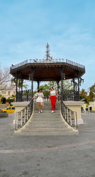 Two women descending the stone steps of an ornate wrought-iron gazebo in a sunny town plaza, with palm trees, colonial-style yellow buildings and bright blue sky.