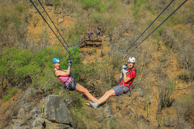Two riders on parallel ziplines over a rocky, cactus-strewn canyon, wearing helmets and harnesses, smiling and touching feet during a zipline adventure