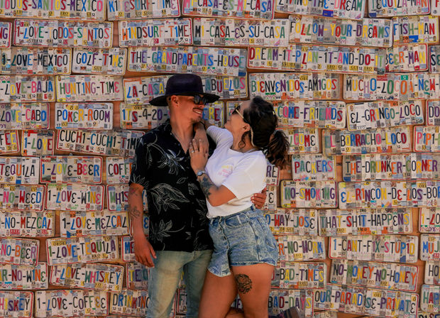 Couple laughing and embracing in front of a colorful Mexican novelty license-plate mural covered in playful phrases — man in a hat and floral shirt, woman in a white tee and denim shorts with a thigh tattoo.