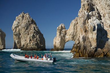 Visitors in orange life jackets waving from an inflatable tour boat passing the iconic El Arco rock arch at Land's End, Cabo San Lucas, with turquoise ocean and clear blue sky.