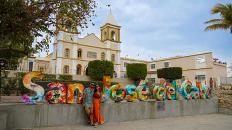 Vibrant 'San José del Cabo' painted letters with two people posing in front of a yellow-and-white mission-style church and manicured trees under a blue sky