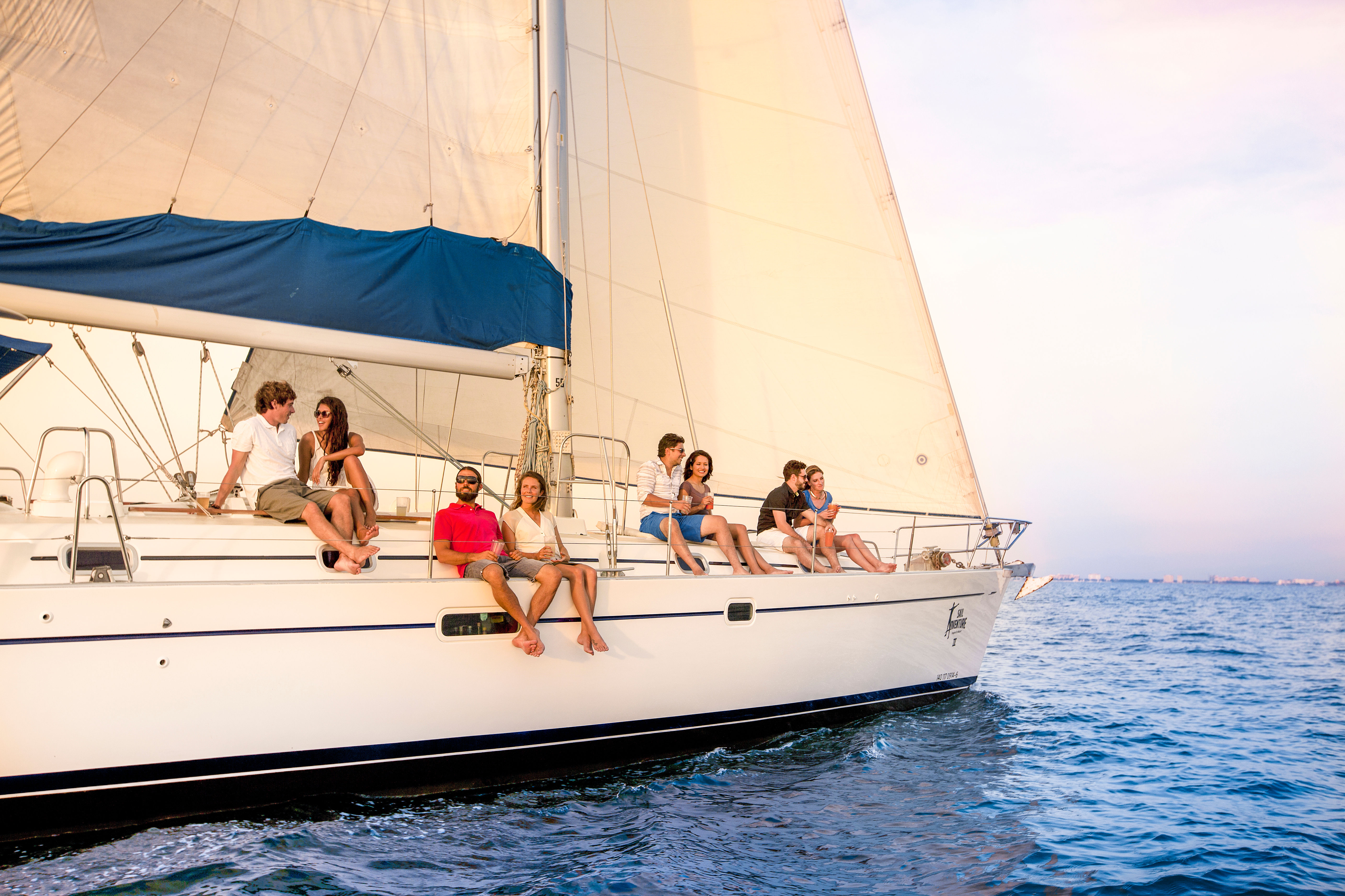 Group of friends lounging on the deck of a white sailboat with billowing sails, enjoying a sunset cruise on calm blue ocean waters