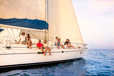 Group of friends lounging on the deck of a white sailboat with billowing sails, enjoying a sunset cruise on calm blue ocean waters