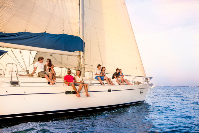 Group of friends lounging on the deck of a white sailboat with billowing sails, enjoying a sunset cruise on calm blue ocean waters