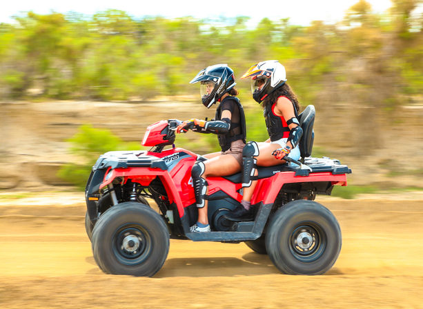 Two helmeted riders on a red ATV (quad) speeding across a dusty off-road trail with blurred desert shrubs in the background.