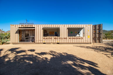 Beige shipping container converted into an outdoor plastic recycling workspace with open service windows showing tools and shelving, set on a sunlit dirt lot under a clear blue sky with distant hills.