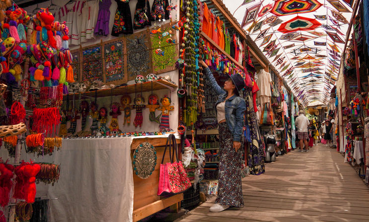 Person browsing a colorful artisan market aisle filled with Mexican-style textiles, tassel garlands, painted skulls and handmade souvenirs under a patterned canopy