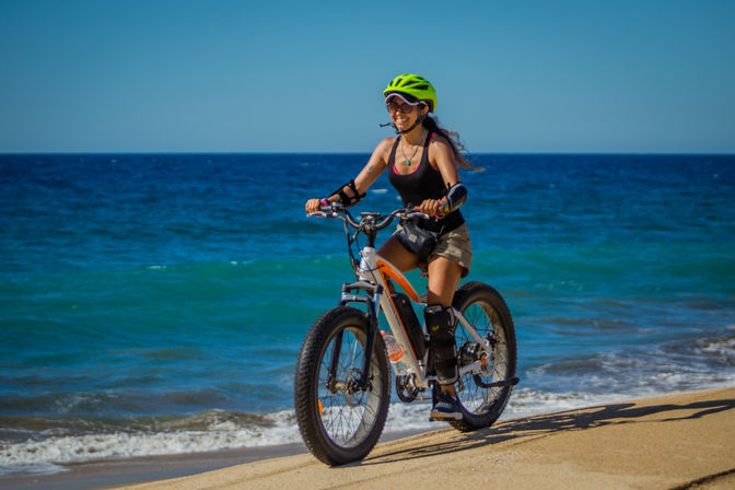 Smiling cyclist in neon green helmet riding a fat-tire bicycle along a sunlit sandy beach with turquoise ocean waves