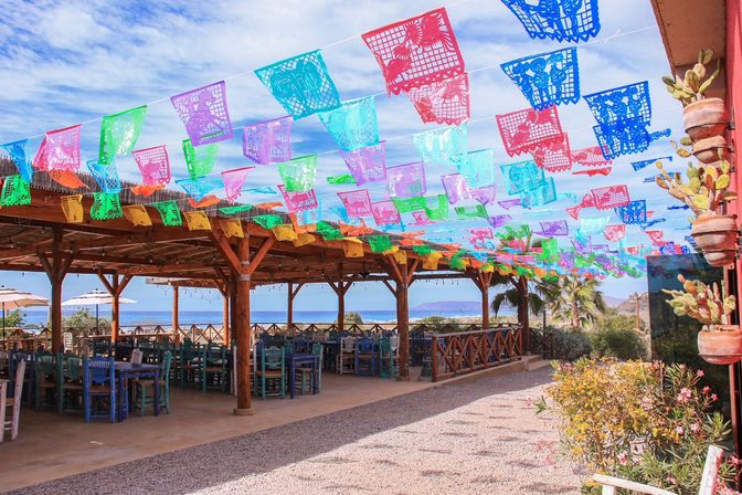 Festive beachside outdoor dining patio with wooden pergola and colorful papel picado banners overhead, turquoise chairs, potted cacti and a clear ocean view