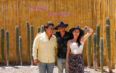 Three smiling friends taking a selfie in front of a bamboo fence lined with tall cacti and a pink neon "TODOS SANTOS" sign, sunny desert vibe.