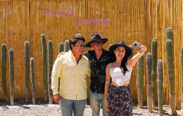 Three smiling friends taking a selfie in front of a bamboo fence lined with tall cacti and a pink neon "TODOS SANTOS" sign, sunny desert vibe.