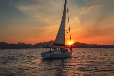 Sailboat on the ocean at golden sunset — passengers enjoying a sunset sail with the sun dipping behind silhouetted coastal hills and warm reflections on the water.
