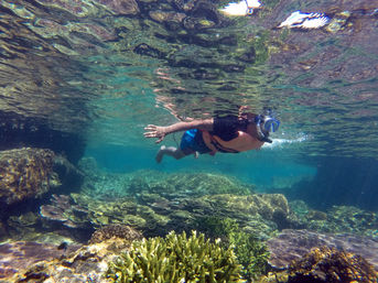 Snorkeler in a blue mask and orange life vest gliding just below the surface over a vibrant tropical coral reef with clear turquoise water and layered plate corals.