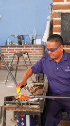 Artisan glassblower wearing safety goggles shapes glowing orange molten glass on a blowpipe in a brick-walled workshop with tools and workbench in the background.