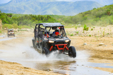 Thrilling off-road UTV splashing through a shallow creek on a sandy desert trail with green foothills and distant mountains.