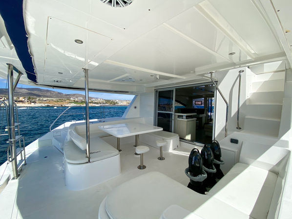 Spacious white luxury yacht aft deck with dining table, bar stools, cushioned seating and sunny ocean coastline view.
