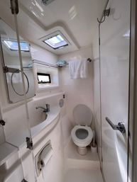 Bright compact boat cabin bathroom (marine head) with white toilet, curved sink and faucet, small porthole window, overhead skylight, towel on rack and mirrored cabinet.