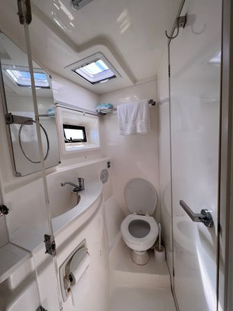 Bright compact boat cabin bathroom (marine head) with white toilet, curved sink and faucet, small porthole window, overhead skylight, towel on rack and mirrored cabinet.