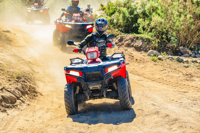 Group of ATV riders on red quad bikes kicking up dust on a sunny off-road dirt trail through scrubby hills, lead rider in helmet and protective gear on an outdoor adventure.