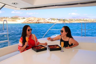 Two women in sunglasses enjoy a yacht brunch with mimosas and bento-style food boxes on a sunny day, blue ocean and rocky coastline visible in the background.