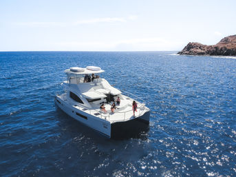 Sunlit luxury catamaran yacht cruising deep-blue ocean near a rocky coastline, guests relaxing on bow and flybridge.