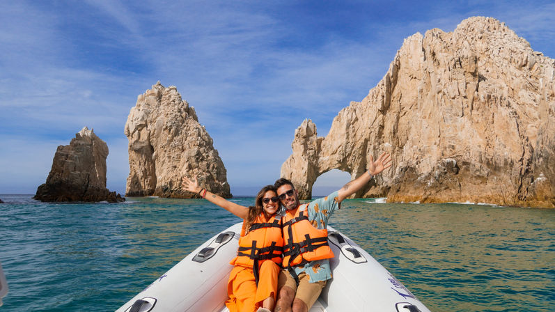 Two people in bright orange life jackets cheering on an inflatable boat in turquoise Sea of Cortez waters with the towering rock arch El Arco at Land's End, Cabo San Lucas, Mexico under a clear blue sky.