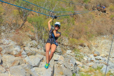 Smiling person in helmet and harness ziplining above a rocky canyon and dry scrubland — outdoor zipline adventure.