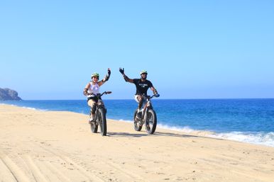 Two cyclists in helmets high-fiving while riding fat-tire e-bikes along a sunny sandy beach beside clear blue ocean waves