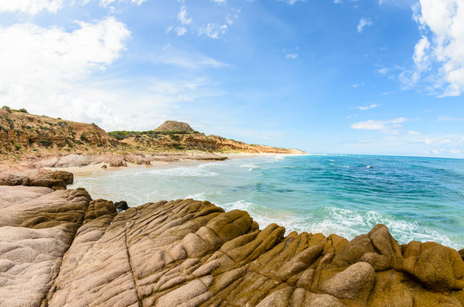 Scenic sunlit rocky coastline with layered sandstone outcrops, a sandy beach and turquoise waves under a bright blue sky.