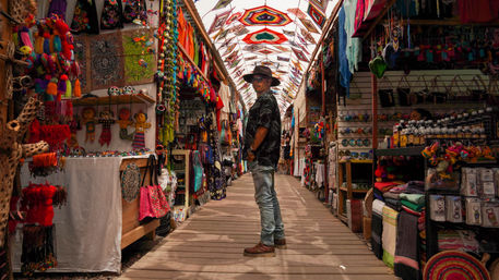 Visitor in a hat standing in a colorful open-air Mexican artisan market aisle lined with handmade textiles, woven bags, dolls, and hanging kites.