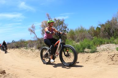 Cyclist in a green helmet and purple tank top riding a white-orange fat‑tire e‑bike on a sandy off‑road trail through low scrub under a bright blue sky, with other riders following in the background.