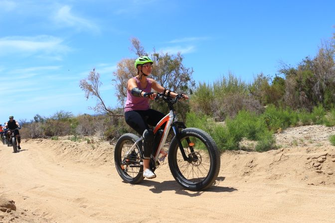 Cyclist in a green helmet and purple tank top riding a white-orange fat‑tire e‑bike on a sandy off‑road trail through low scrub under a bright blue sky, with other riders following in the background.