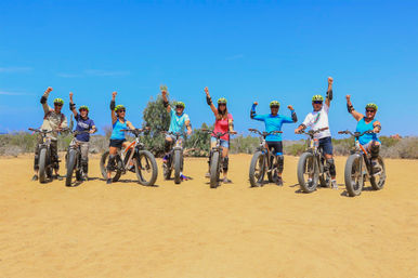 Eight riders on fat-tire e-bikes on a sunny sandy desert trail, wearing helmets and pads and cheering with raised fists under a bright blue sky.
