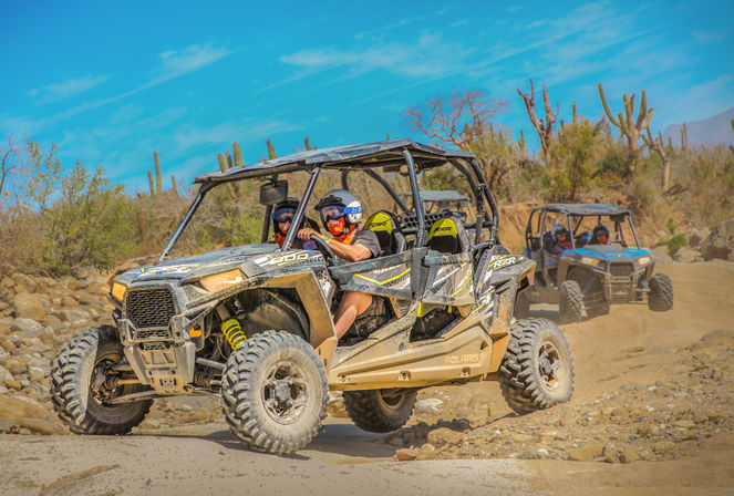 Dusty desert trail with two off-road side-by-side UTVs kicking up dust, drivers in helmets and goggles, rocky path and cactus-studded landscape.