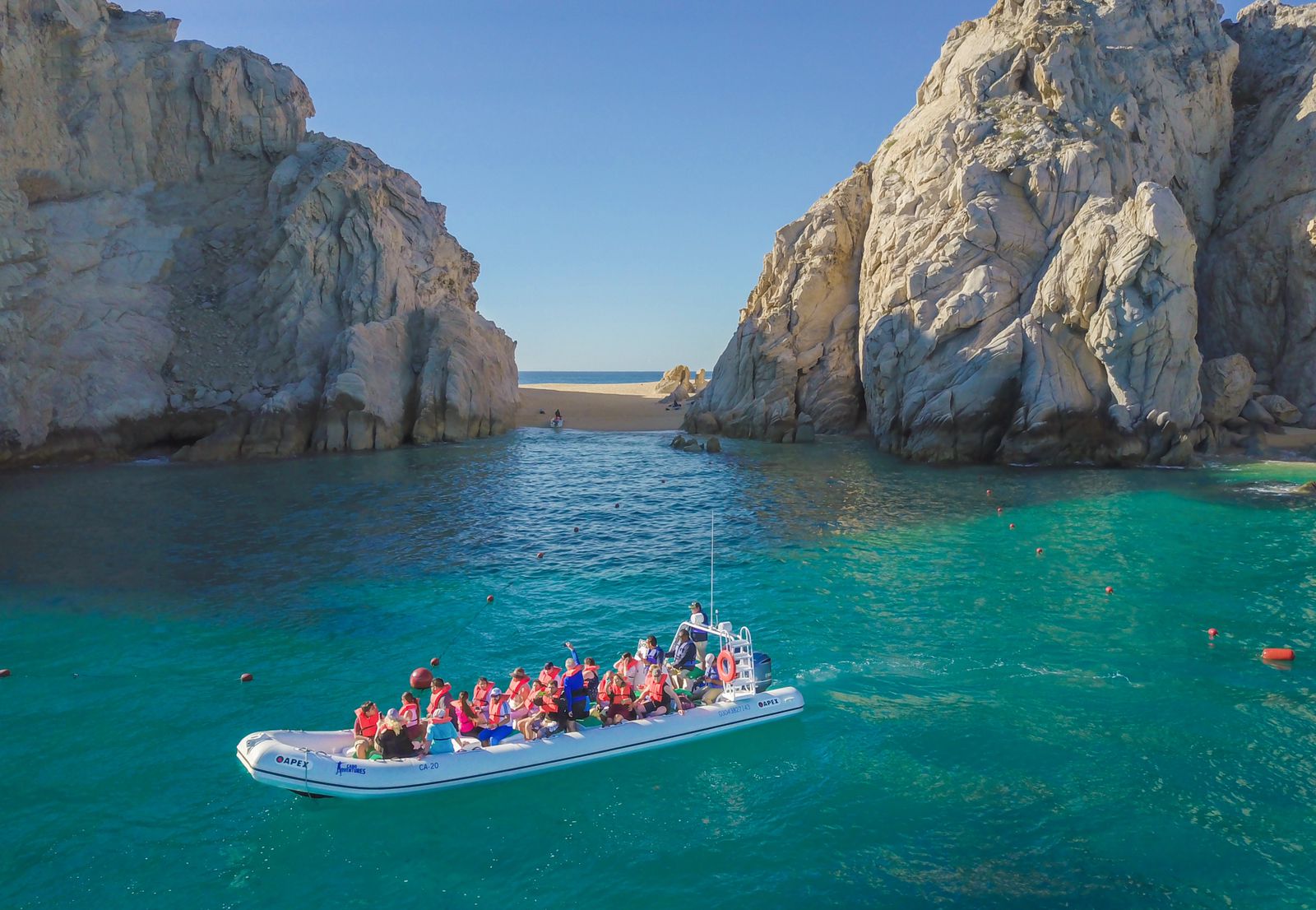 Tour boat full of passengers in life jackets on turquoise water approaching a sandy cove framed by towering limestone cliffs under a clear blue sky